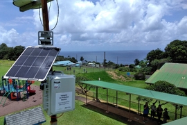 Photo of a solar-powered air quality sensor, labeled with a sticker that says "volcanic air pollution monitor," installed next to a playground and park next to the ocean