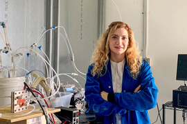 Photo of a woman wearing a blue lab coat, her arms folded, leaning against a workbench containing research equipment.