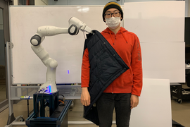 Photo of a masked student standing in front of a white background while a robot arm helps him put on a jacket.