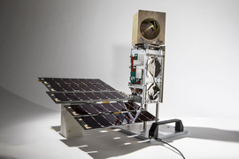 Photo of a small satellite against a white backdrop. The satellite has solar cells toward the bottom and a small boom with a gold cube at the top.