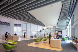 Photo of the interior of Hayden Library with seating areas and two people standing at a desk in the foreground; in the background is a glass-enclosed room and a staircase.