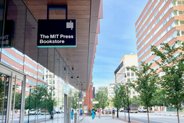Photo of a side view of a glass-covered building, at left, with a black sign with "The MIT Press Bookstore" hanging from an overhang. The photo looks west with other buildings across the street and short trees along the sidewalk.