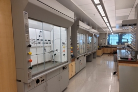 Photo showing some fume hoods in a lab. The hoods are enclosed work stations, several feet across and several feet wide, with pipes and other equipment inside of a glass window