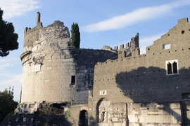 Photo of castle ruins. At left is a circular tower partially destroyed and missing a roof. At right is a partially intact facade of brick, also missing a roof, with columns and ancient sculptures embedded toward the bottom.