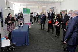 Photo of six people in suits and face masks watching a presentation by a young woman, who is laughing and who is demonstrating with a small spherical robot