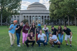 Photo of 12 young people in summer clothes posing before MIT’s Great Dome, each pointing with their index fingers. Most are pointing up, two in other directions, and one not at all.