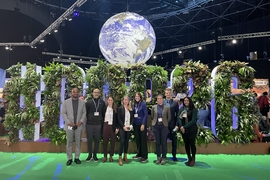 Photo of 8 MIT affiliates stand in a large convention center, in front of a sign made with leaves that reads “#COP26.” A large Earth globe hangs down behind them.