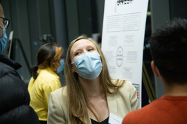 Photo of a woman wearing a mask, standing in front of a poster