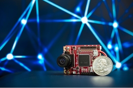 Photo of a small brown circuit board with a camera attached, next to a US 25-cent coin for scale.
