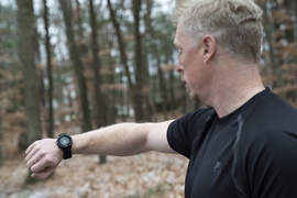 Photo of James Balcius looking at the smartwatch on his wrist. The small circular watch display shows a green circle with a thumbs-up sign in the middle. He is outside, with trees and fallen leaves in the background.