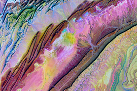 An aerial view of mountains in iridescent shades of pinks, greens, yellows and browns. The colors fold and spill against each other.