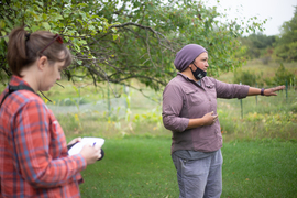 Photo of ESI Journalism Fellow Nora Hertel standing in a grassy field next to a tree, taking notes while Jessika Greendeer looks and points to the right