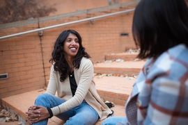 Photo of Athulya Aravind sitting on some steps and smiling while conversing with an unidentified companion
