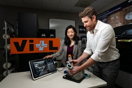 Two researchers standing at a table inside of a lab showing the AI-Guide handheld device and an ultrasound display.