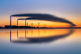 three smokestacks billow symmetric plumes of smoke over the horizon, which is reflected in a pool of still water below.