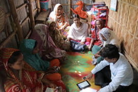 In Bangladesh, a group of eight women sit in a circle in a small room. A man, also sitting on the floor with them, is explaining a BRAC humanitarian program. An iPad and cell phone are on the floor. 
