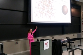 Polina Anikeeva stands behind a podium pointing to a slide on a screen behind her that is front of a blackboard