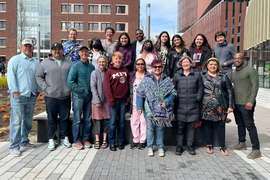 Photo of a group of 19 smiling people posing in a group photo in front of several MIT buildings