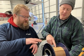 Ben Evans and Dave Whelihan point to and discuss a large spool of copper wire. They are standing inside a hangar with a small airplane behind them. 