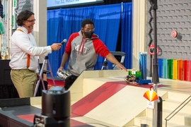 Photo of Brandon Worrell pointing to a robot as Amos Winter interviews him. They are next to a colorful robot obstacle course.