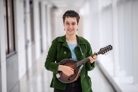 Photo of Zoe Levitt standing in the center of the frame in a white hallway, holding a mandoline and smiling.