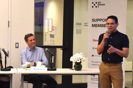 One person on the right stands, speaking with a microphone in hand; another person on the left is seated at a table, listening.