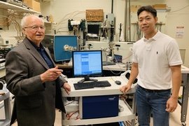 Harry Tuller and student pose for a photo in a lab, with a computer screen on a table between them showing data