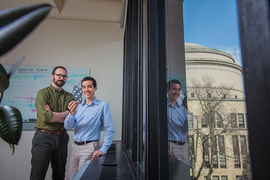 Photo of Rafael Gómez-Bombarelli and Daniel Schwalbe-Koda standing together in an office next to a window through which you can see the MIT dome. Schwalbe-Koda holds up a zeolite model.