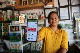 A retail shop owner in India holds a cell phone to the camera showing the Essmart interface.