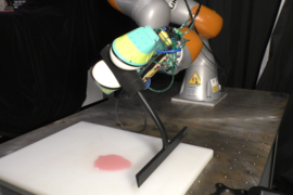 Photo of a robotic arm holding a squeegee in preparation to wipe up a blob of pink liquid