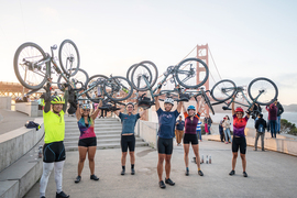 Six bicyclists in full riding gear stand in front of the Golden Gate Bridge holding their bicycles above their heads in triumph