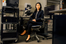 Gloria Choi sits on a chair surrounded by grey and black equipment in a lab, including a microscope, computer monitors, and rolling carts with devices on them. 