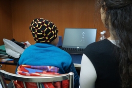 Photo from behind a person sitting at a desk, watching dot patterns on a monitor, while another person looks on. 