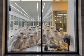 A large group of students, wearing full-body white lab suits, posing in the window of the MIT.nano cleanroom, seen from the outside.