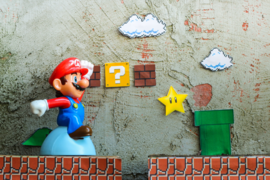 A figurine of video game character Mario runs across a cardboard cutout environment, which features several brown pixelated bricks, a yellow mystery box, a magical yellow star, and a green pipe.