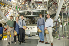 Five scientists, each holding a gold-colored hardhat, stand in front of a huge machine known as magnetic recoil neutron spectrometer, designed at MIT