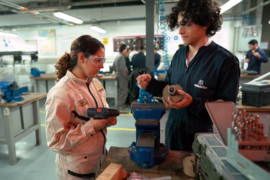 Two students in a workshop wearing safety goggles and jumpsuits. The student on the left is holding a drill. The student on the right is holding materials for the project on the table between them.