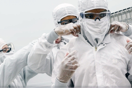 In a clean room, 3 people wear full-body clean suits. They help zip up each other’s suits.