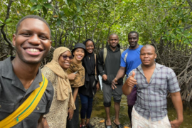 Seven individuals stand and pose for a group photo in a lush green wooded area.