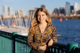 Aviva Intveld, smiling and leaning on a railing overlooking the Charles River with the Boston skyline in the background