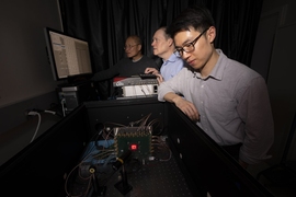 Three men stand at a lab bench in a dark room. Two are looking at a computer screen and the third is examining a circuit board festooned with wires.