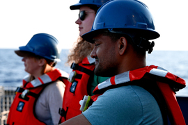 Three people wearing life jackets and hard hats look at the ocean aboard a boat.