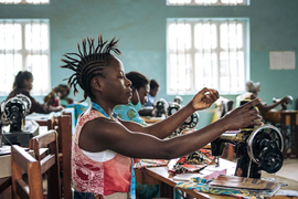 A woman with a mohawk-style braided hairstyle works at a sewing machine with brightly-colored fabrics, alongside other women