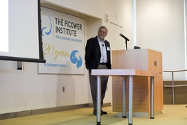 Mriganka Sur stands at a lectern in an auditorium. Behind him is a banner with the Picower Institute's 20th Anniversary logo.
