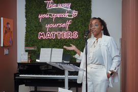 Lauryn McNair stands, speaking into a microphone. On the wall behind her is a pink neon sign atop fake grass that says "The way you speak to yourself MATTERS." A piano sits below that.