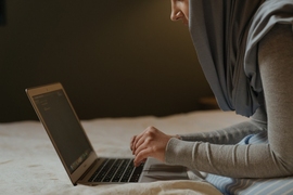 Photo of a woman wearing a hijab working on a laptop. The photo is cropped such that you can only see from her nose down, with the hajib covering most of the side of her face