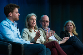 Four panelists sit on a stage. One is speaking and gesturing while the other three watch.