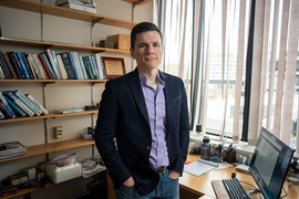 Robert Macfarlane stands in an office, a desk in front of him, bookshelves behind him, and a wall of windows with vertical blinds.