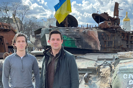 Ian Miller and Evan Platt stand in front of a battered tank. A Ukranian flag flies from the tank.