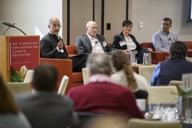 Four adults sit in red armchairs beside a podium bearing a sign that reads, MIT Symposium for Advancing Climate Education, April 2023. Man at the far left gestures toward the seated audience.